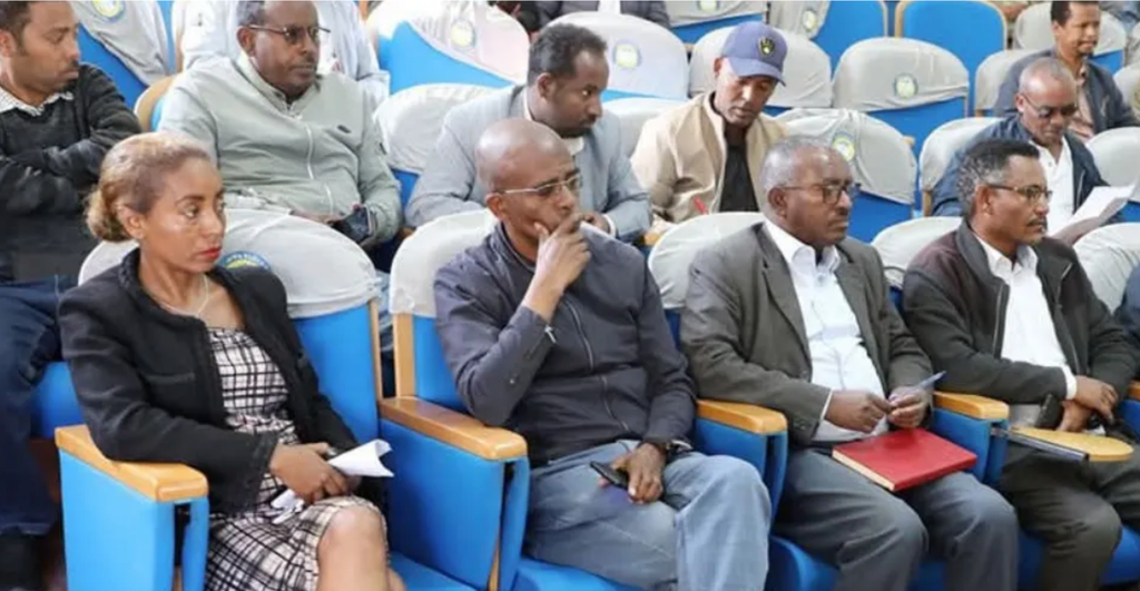 Participants seated in an auditorium listen attentively during a session, some holding notes and folders.