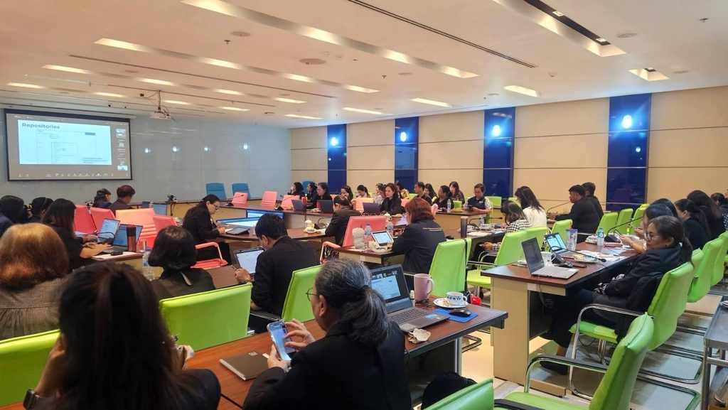 A workshop in a bright conference room, with participants working on laptops at long tables while a slide is projected on a screen at the front.